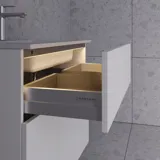 Stylish bathroom inspiration with beige vanity unit with inner drawer, upper cabinet, open shelf, oval mirror with lighting, matching pendants, shelf under the mirror, and wall-mounted toilet. Walls with light, elongated tiles.